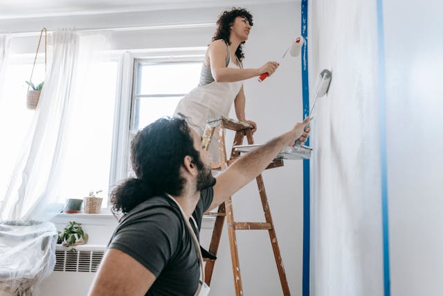 two-people-painting-a-wall-white-one-standing-on-a-ladder-and-the-other-reaching-up-with-a-roller-in-a-bright-room-with-natural-light two-people-painting-a-wall-white-one-standing-on-a-ladder-and-the-other-reaching-up-with-a-roller-in-a-bright-room-with-natural-light